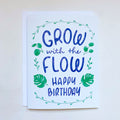 A greeting card with 'Grow with the Flow Happy Birthday' in blue letters, surrounded by illustrations of plant leaves, monstera plant. The card is placed against a light background. It has a white envelope