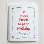 Greeting card with red text 'sending love on your birthday' and green leaves on a beige background