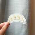 Hand holding a cartoon-style dumpling sticker against a gray background