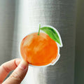 Hand holding a sticker of an orange with a green leaf against a metallic surface