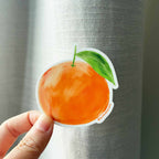 Hand holding a sticker of an orange with a green leaf against a metallic surface